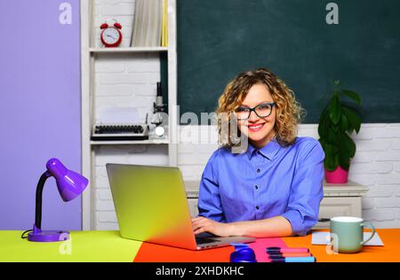 Professeur d'école souriant ou tuteur d'université assis au bureau dans la salle de classe. Retour à l'école. Professeur d'université travaille avec un ordinateur portable. Étudiante préparant fo Banque D'Images