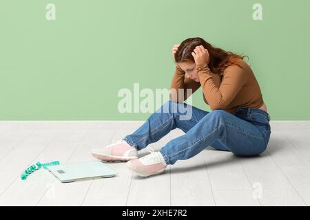 Femme en surpoids troublée en jeans serrés avec des écailles et du ruban à mesurer sur fond vert. Concept d'excès de poids Banque D'Images