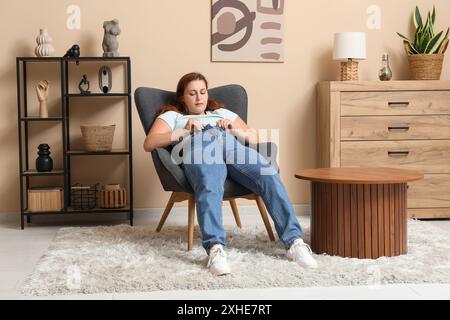 Femme en surpoids en jeans serrés assis sur un fauteuil à la maison. Concept d'excès de poids Banque D'Images