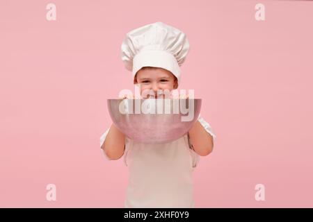 Enfant heureux, habillé en chef avec un chapeau et un tablier, tient un grand bol pour un portrait dans un cadre de studio rose. Enfant âgé de 3 ans (garçon de 3 ans) Banque D'Images