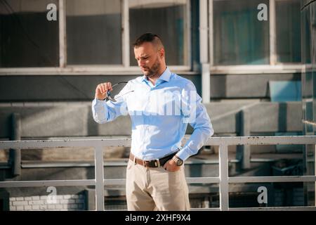 Un homme dans une chemise bleu clair et un pantalon beige se tient sur un toit, ajustant ses lunettes. Il est devant une balustrade blanche et un bâtiment avec grand Banque D'Images