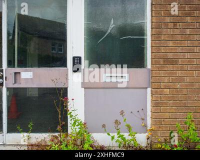 Bâtiment abandonné avec des plantes envahies près d'une vieille porte vitrée, avec un clavier et une fente de courrier. Banque D'Images