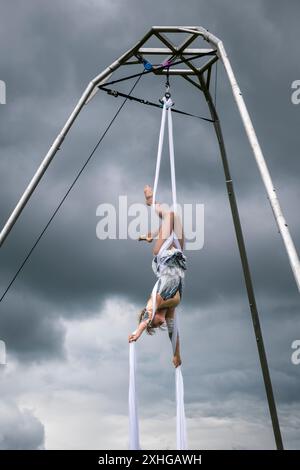 Une acrobate jouant des soies aériennes à l'extérieur sous un ciel orageux lors d'un carnaval local, pendant le mauvais été de 2024 (Royaume-Uni) Banque D'Images