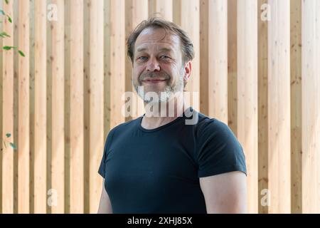 Un homme âgé avec une barbe sourit à la caméra, debout devant un mur de bois. Banque D'Images