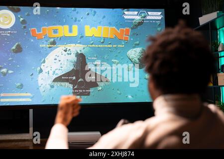 Homme afro-américain jouant à des jeux vidéo sur console de jeu, célébrant la victoire. Gamer excité après avoir gagné un match de jeu multijoueur en ligne sur l'écran ultra-large Smart TV, se réjouissant Banque D'Images