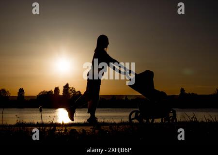 Silhouette d'une mère tirant une poussette contre le coucher du soleil près du lac. Banque D'Images