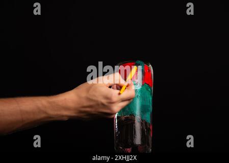 Une main de peintre peint une bouteille en verre avec des peintures. Isolé sur fond noir. Banque D'Images