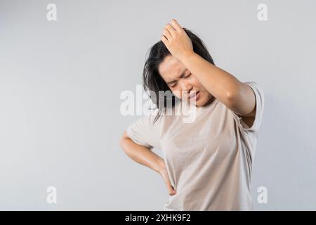 Femme asiatique frustrée tient les mains sur la tête, a mal à la tête, semble misérable, ne peut pas gérer la pression, se sent en détresse, se tient sur fond gris. Menta Banque D'Images