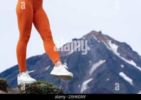 Femme méconnaissable faisant de l'entraînement de fitness dans les montagnes avec volcan en arrière-plan. Gros plan des jambes dans la silhouette orange serrant les leggings de sport et Whit Banque D'Images