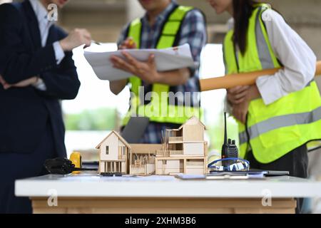 Modèle architectural d'une maison avec plan et outils sur table, équipe d'ingénieurs travaillant en arrière-plan flou. Banque D'Images