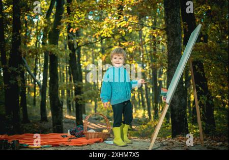 Petit artiste peignant tableau. Amitié d'enfance et premiers souvenirs. Camping avec enfants. Petit garçon en pull ou pull bleu vintage tendance. Banque D'Images