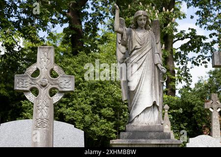 Une croix celtique à côté d'une statue en pierre d'un ange avec un fond d'arbres et de feuillage, dans le cimetière de Highgate, Londres, Royaume-Uni Banque D'Images