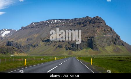 Paysage montagneux du sud de l'Islande avec route périphérique droite par jour ensoleillé avec des prairies vertes et des montagnes rocheuses enneigées en arrière-plan. Banque D'Images