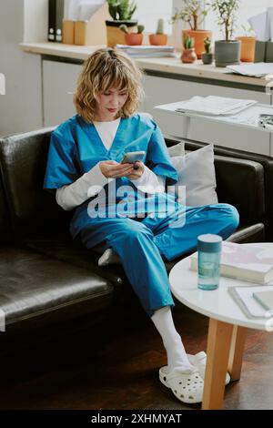 Jeune infirmière caucasienne vêtue d'un uniforme médical bleu vérifiant les réseaux sociaux tout en étant assise sur un canapé en cuir noir dans la salle d'appel pendant sa pause Banque D'Images