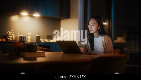Belle femme asiatique authentique assise à une table dans Cozy Kitchen et à l'aide d'un ordinateur portable à la maison la nuit. Femmes souriant et discutant sur Internet et les médias sociaux, faisant de la recherche scientifique. Banque D'Images