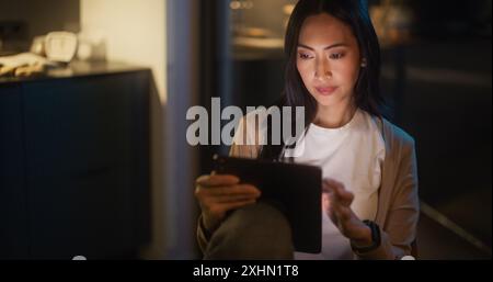 Belle femme asiatique authentique assise à une table dans Cozy Kitchen et utilisant la tablette numérique à la maison la nuit. Femme souriant et vérifiant les médias sociaux, faire des achats en ligne, regarder des vidéos Internet Banque D'Images