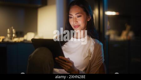 Belle femme asiatique authentique assise à une table dans Cozy Kitchen et utilisant la tablette numérique à la maison la nuit. Femme souriant et discutant sur les médias sociaux, faisant des achats en ligne, naviguant sur Internet Banque D'Images