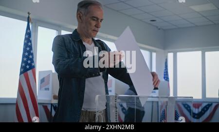 Divers électeurs, les Américains votent dans les cabines de vote au bureau de vote. Un homme caucasien adulte met un bulletin de vote dans une boîte. Jour des élections nationales aux États-Unis d'Amérique. Devoir civique et concept patriotique. Banque D'Images