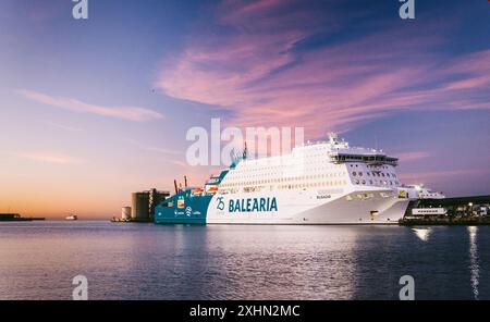 Profil coucher de soleil du ferry BALEARIA amarré dans le port animé, eaux sereines reflétant les teintes du ciel rosé. Structures industrielles de fond, deuxième navire Banque D'Images