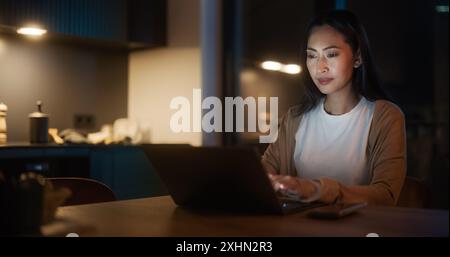 Belle femme asiatique authentique assise à une table dans une cuisine confortable et tapant sur ordinateur portable à la maison la nuit. Femme souriant et discutant sur les médias sociaux, faisant des achats en ligne, vérifiant les nouvelles. Banque D'Images