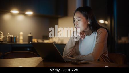 Belle femme asiatique authentique assise à une table dans une cuisine élégante et utilisant un ordinateur portable à la maison la nuit. Femme souriant et discutant sur les médias sociaux, faisant des achats en ligne, vérifiant les nouvelles. Banque D'Images
