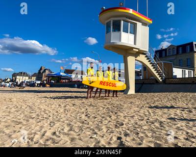 Lion-sur-mer, France, 14 juillet 2024. Les sauveteurs de mer se tiennent sur la plage en T-shirts jaunes avec un équipement de sauvetage. A côté du stand il y a une baguette Banque D'Images