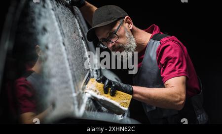 Spécialiste professionnel en lavage de voiture étalant de la mousse avec une éponge souple pour préparer un SUV électrique noir moderne au design contemporain dans un centre de voiture de concession. Commercial Studio photo pour la publicité Banque D'Images