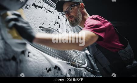 Portrait d'un homme adulte travaillant dans un studio d'esthétique, préparant un VUS électrique frais d'usine pour les travaux d'entretien et le traitement d'entretien de voiture. Technicien de nettoyage utilisant une éponge avec de l'eau savonneuse Banque D'Images