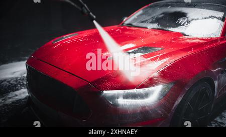 Expert en lavage de voiture utilisant une laveuse à pression d'eau pour rincer une voiture de sport moderne rouge. Homme adulte lavant la saleté, préparant une voiture de muscle américaine pour le détail. Photo cinématographique créative avec véhicule sport Banque D'Images