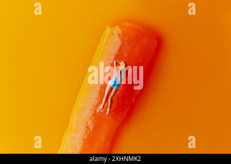 gros plan d'une femme blonde miniature, dans un maillot de bain bleu, couchée face vers le bas sur un gel pop aromatisé orange placé sur un fond orange Banque D'Images