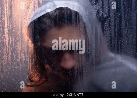 Portrait en gros plan d'une jeune femme portant un imperméable en plastique transparent derrière du verre. Prise de vue en studio. Isolé sur fond sombre. Banque D'Images