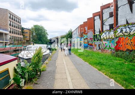 Les gens marchent le long du chemin de remorquage le long du Grand Union canal, Paddington Arm devant des bateaux amarrés et un mur de briques couvert de graffitis Banque D'Images