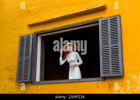 Jolie jeune femme vietnamienne portant une robe traditionnelle Ao Dai et un chapeau conique regardant par la fenêtre avec des volets à Hoi an, Centre du Vietnam, Asie Banque D'Images