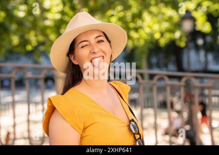 Femme dans un chapeau de soleil et une robe jaune faisant un clin d'œil et tirant sa langue dehors de manière ludique alors qu'à l'extérieur. L'arrière-plan présente de la verdure, suggérant un parc ou un jardin. Banque D'Images