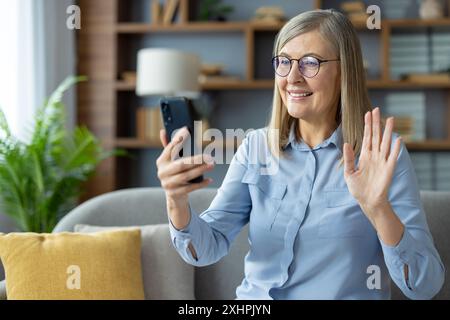 Femme âgée souriante portant des lunettes et une chemise bleue, en utilisant un smartphone pour un appel vidéo. Assis sur le canapé dans le salon confortable, agitant la main et profitant de la communication virtuelle. Banque D'Images