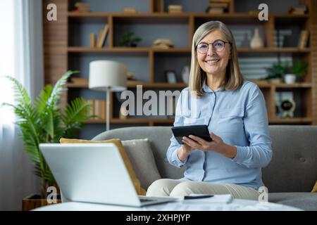 Femme plus âgée utilise la calculatrice pour calculer les factures de services publics et le loyer tout en étant assis confortablement sur le canapé dans la maison moderne avec ordinateur portable et documents. Concept de finances à la maison, budgétisation, et vie à la retraite. Banque D'Images