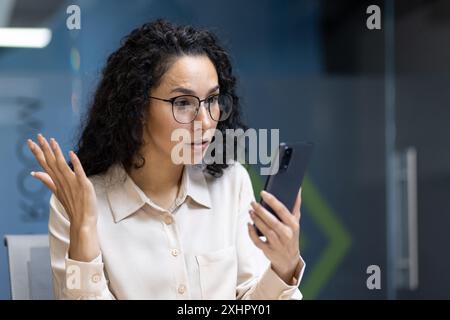 Femme d'affaires avec les cheveux bouclés exprime la surprise et la frustration tout en regardant smartphone dans le bureau. Jeune professionnel assis dans un espace de travail moderne confronté à des défis de travail et de stress. Banque D'Images
