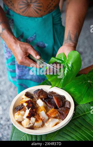 Polynésie française, île de Tahiti, homme polynésien tatoué coupant une banane plantain avec un couteau sur une feuille et la servant dans une assiette avec du taro Banque D'Images