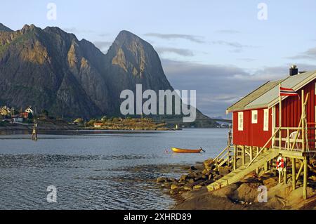 Cabane en bois rouge avec bateau sur la rive du fjord, entouré de montagnes au coucher du soleil, Rorbuer, logement de vacances, Reine, Moskenes, Lofoten, Nordland Banque D'Images