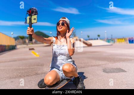 Jeune femme asiatique font du livestreaming joyeusement dans le parc urbain de skateboard agitant à la caméra tout en enregistrant Banque D'Images