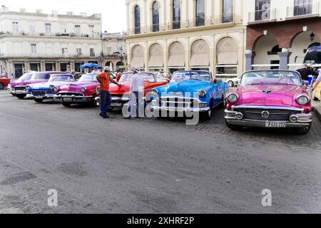 Rangée de cabriolets vintage en rose, bleu et rouge garés dans une rue, les hommes discutant à côté d'eux, Cuba, les grandes Antilles, les Caraïbes, l'Amérique centrale Banque D'Images