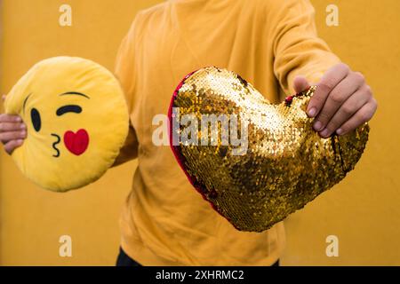 Une personne tenant un oreiller de paillettes en forme de coeur et un oreiller emoji claquant sur un fond jaune Banque D'Images