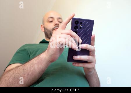 Homme utilisant un téléphone portable haut de gamme, regardant concentré sur l'appareil, portant un polo vert foncé, vu d'en bas, fond blanc Banque D'Images