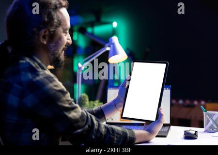 Homme barbu assis au bureau, saisissant une tablette montrant un écran blanc isolé avec un ordinateur portable en arrière-plan. Freelance masculine utilisant un appareil numérique affiche un modèle de maquette vierge. Banque D'Images