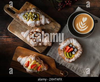 Croissants gastronomiques fraîchement cuits et pâtisserie feuilletée avec baies, confiture, chocolat et pistache. Tasse de cappuccino sur fond en bois foncé. Vue de dessus. Banque D'Images