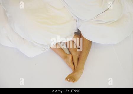 Les pieds d'un couple entrelacés sous des draps blancs confortables, capturant un moment d'intimité, de chaleur et d'amour. La scène respire le confort, la sérénité et la joie Banque D'Images