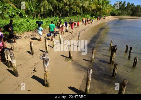 Les villageois portent les bagages des touristes arrivant par bateau, alors qu'ils marchent le long de la plage dans le village de Horale, Seram, Maluku Central, Maluku, Indonésie. Banque D'Images