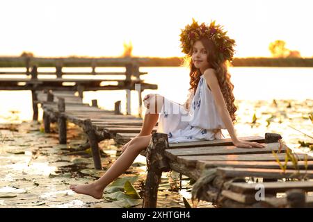 Mignonne petite fille portant une couronne faite de belles fleurs sur la jetée près de la rivière au coucher du soleil Banque D'Images