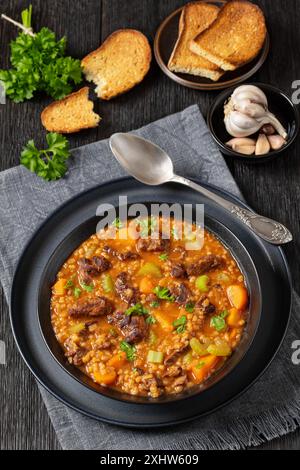 soupe classique à l'orge de bœuf avec de tendres morceaux de rôti de bœuf, des légumes frais et un bouillon assaisonné dans un bol noir sur une table en bois de chêne foncé avec du pain et une cuillère Banque D'Images