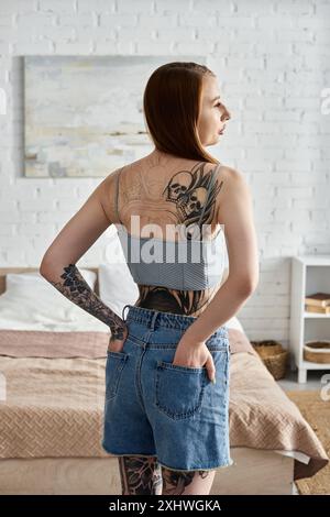 Une jeune femme avec des tatouages sur le dos et les bras se tient dans son appartement moderne, les mains dans ses poches, regardant par la fenêtre. Banque D'Images